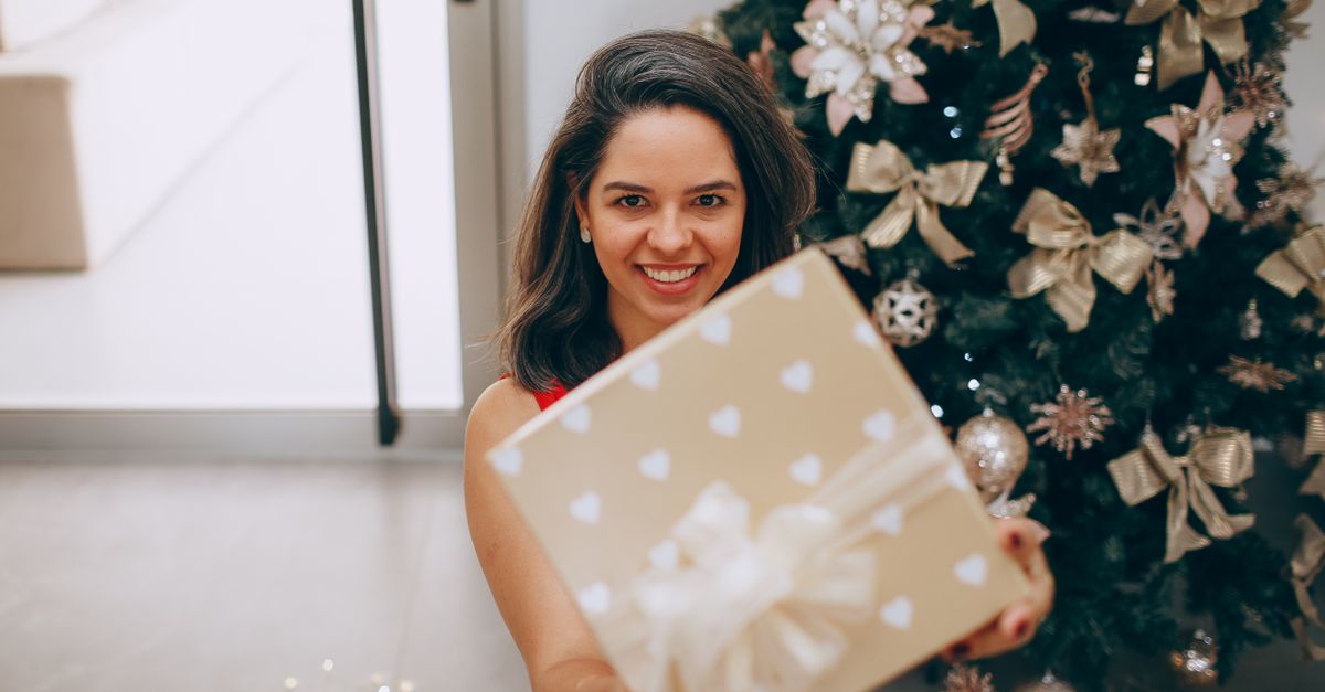 Smiling woman holding a Christmas gift in front of a decorated tree indoors.