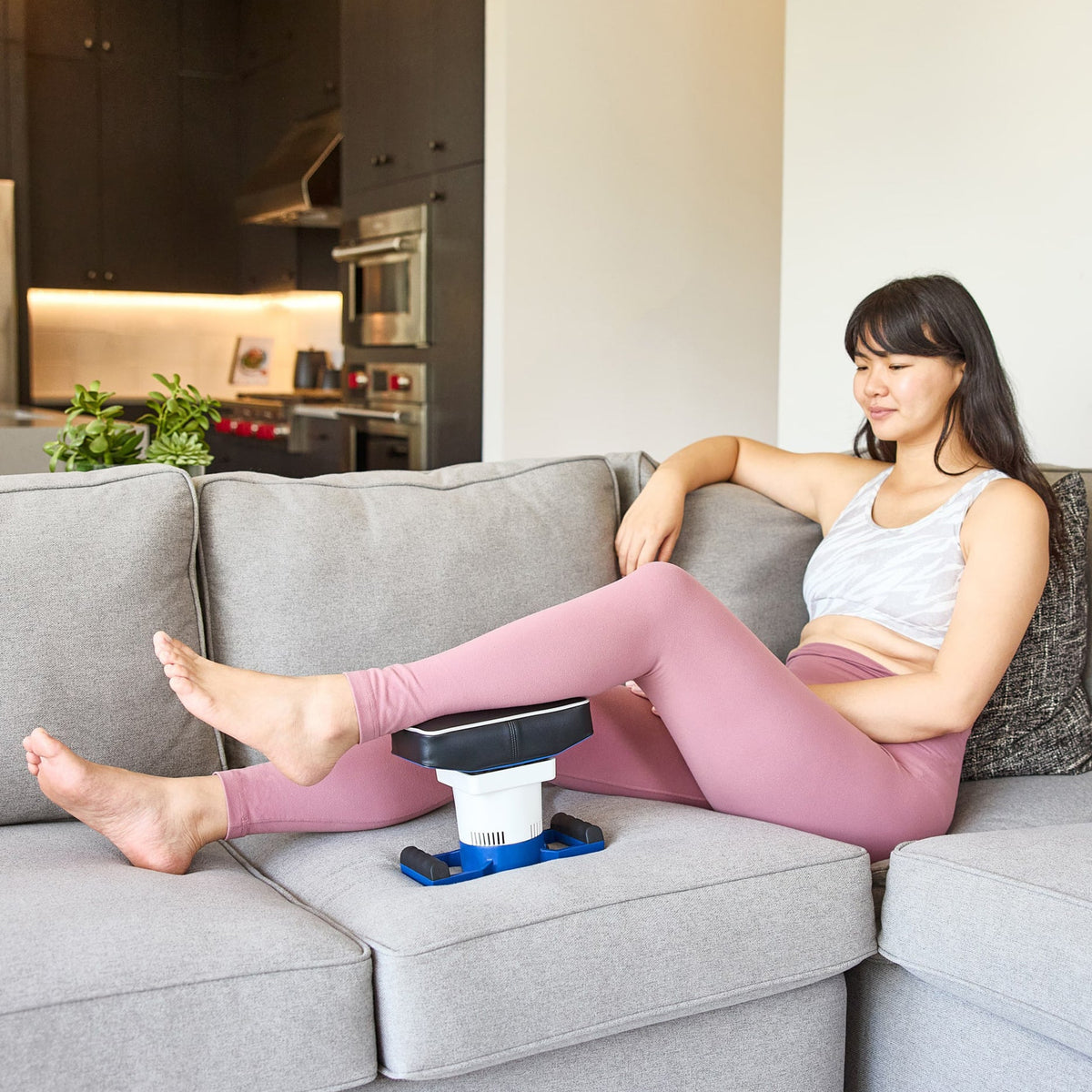 Woman in workout gear relaxing on couch using MedMassager body massager for back and thigh relief.