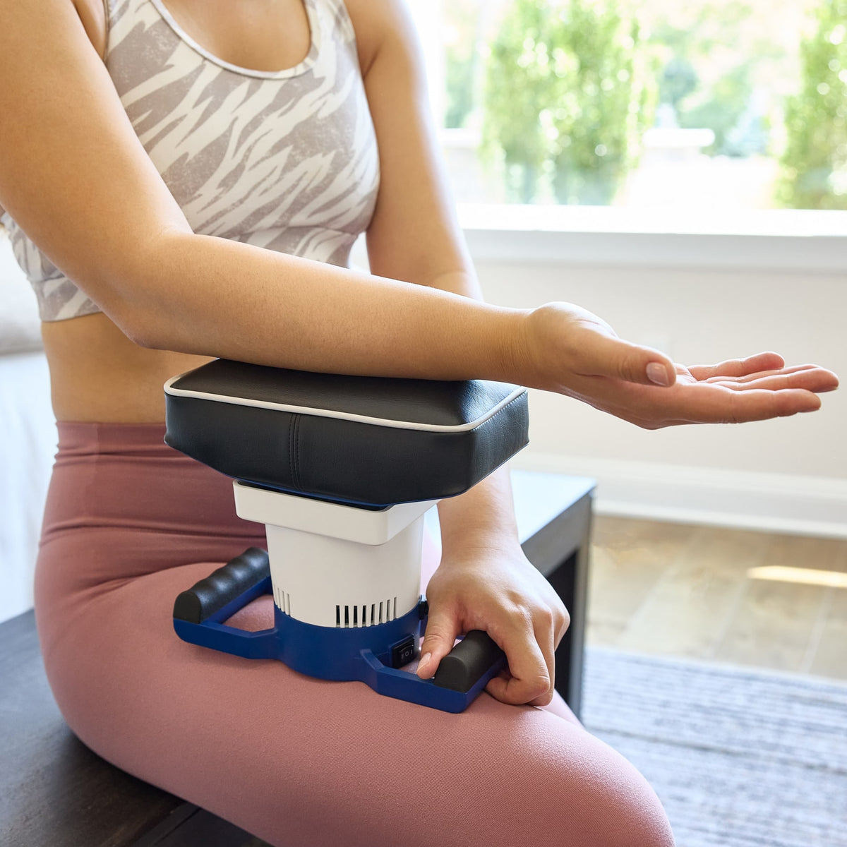 Person in athletic wear uses a MedMassager body massager on their forearm while sitting indoors.