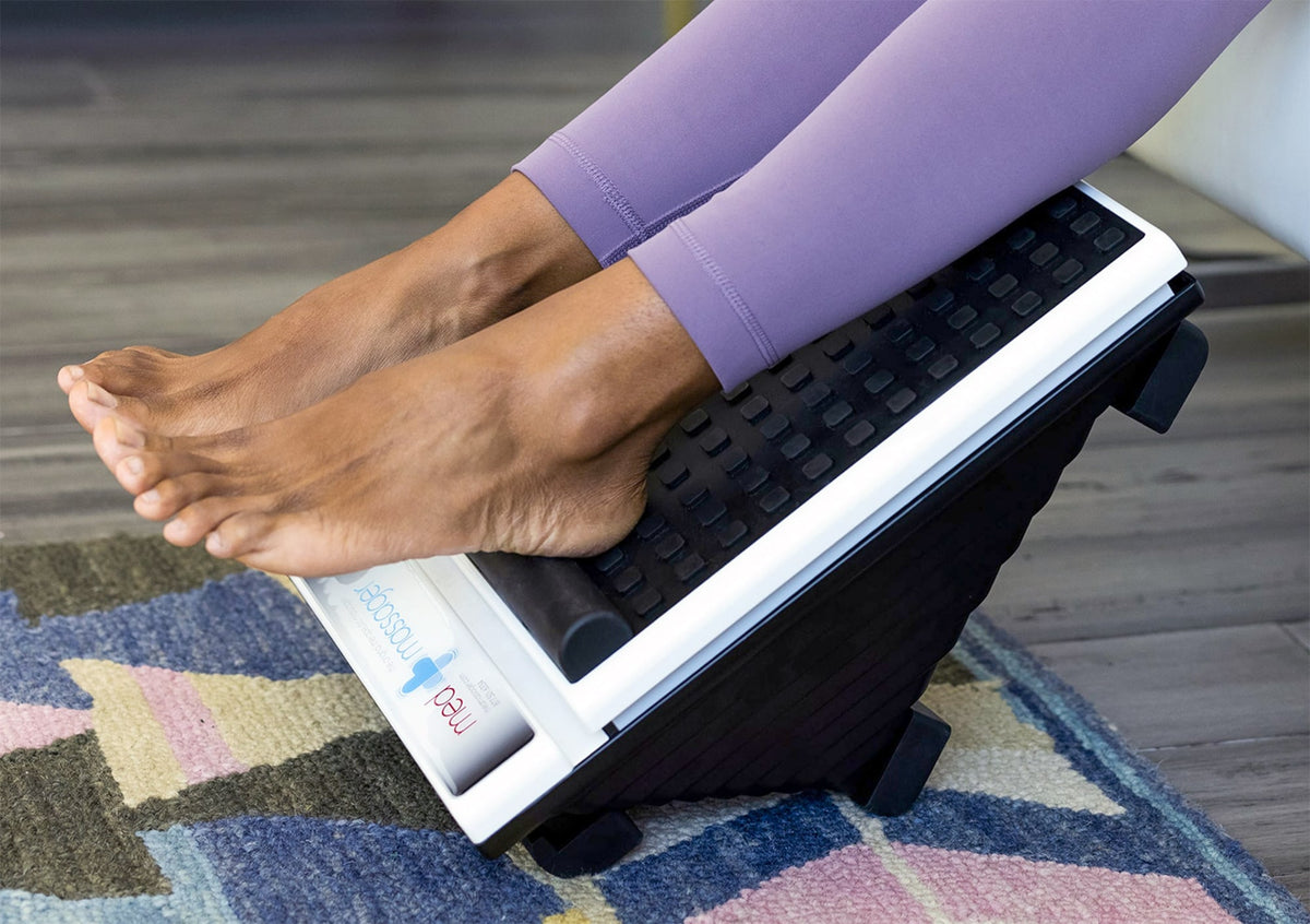 Bare feet on a black and white foot massager, user seated on a colorful rug. MedMassager device in home use setting.