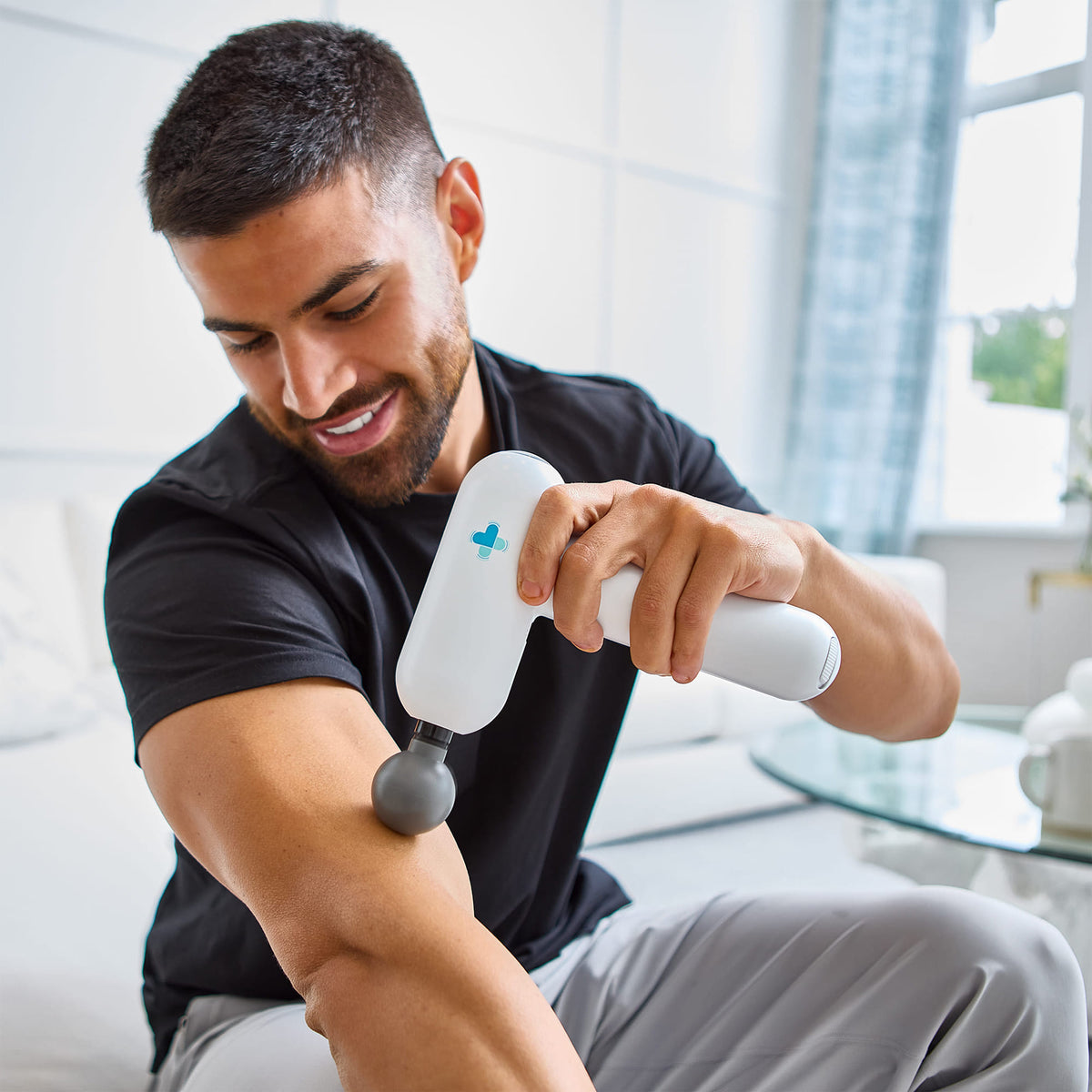 Man using MedMassager body massager on arm for percussive therapy while sitting on a couch.