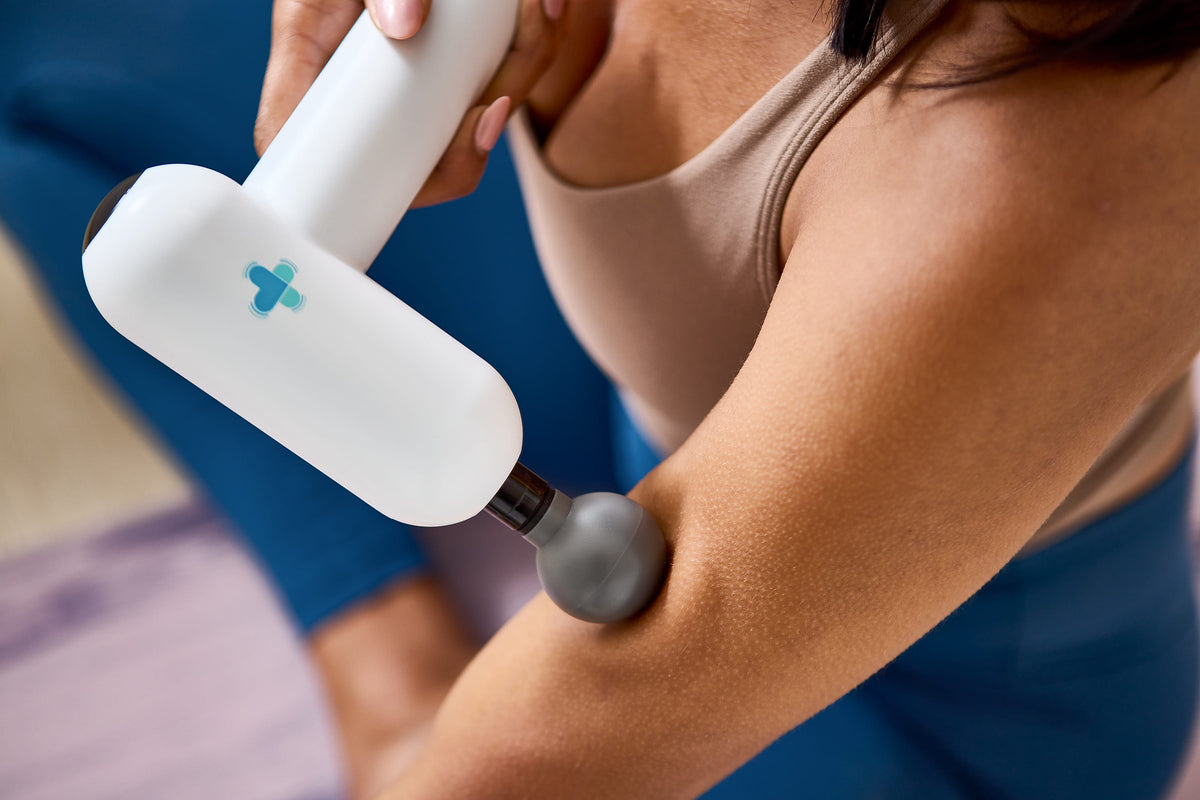 Person using a white MedMassager body massager on their arm, wearing a sleeveless top and blue leggings.