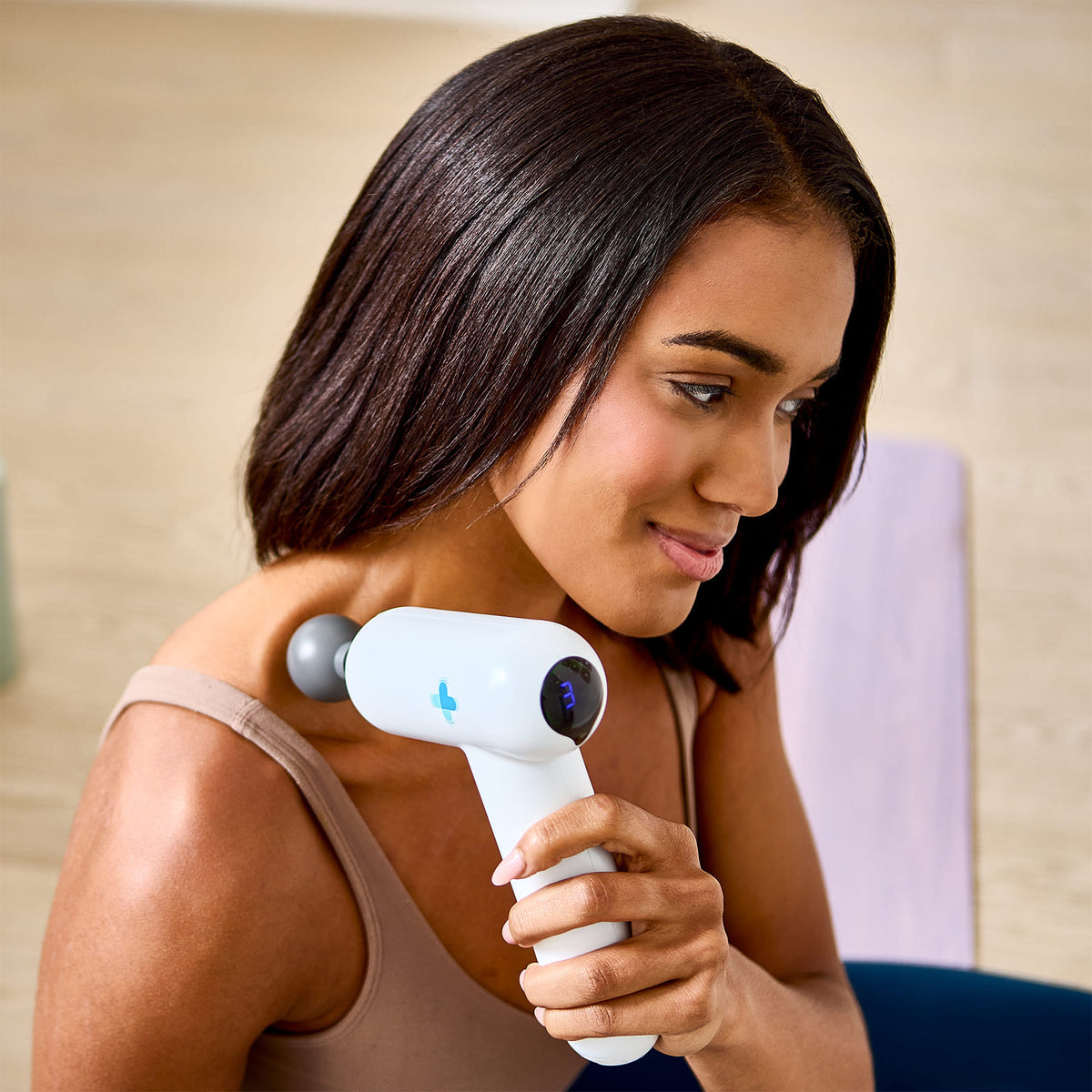 Woman using MedMassager body massager on her shoulders indoors, sitting on a light floor.