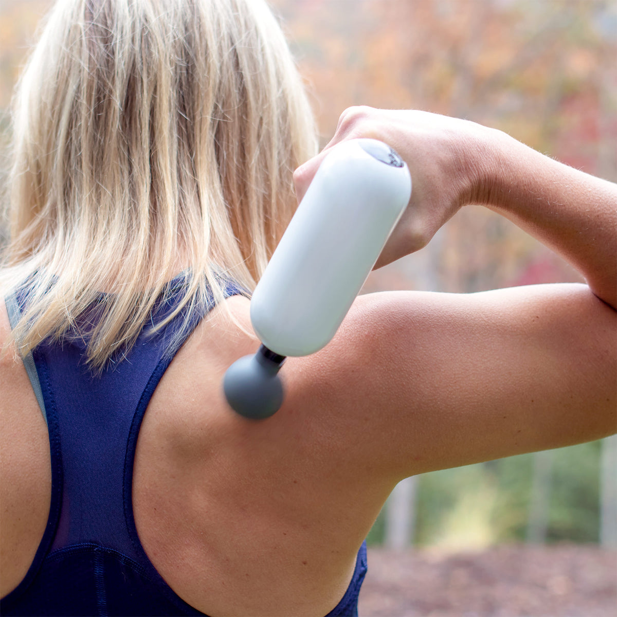 Woman using MedMassager body massager on her upper back outdoors, wearing a blue tank top.