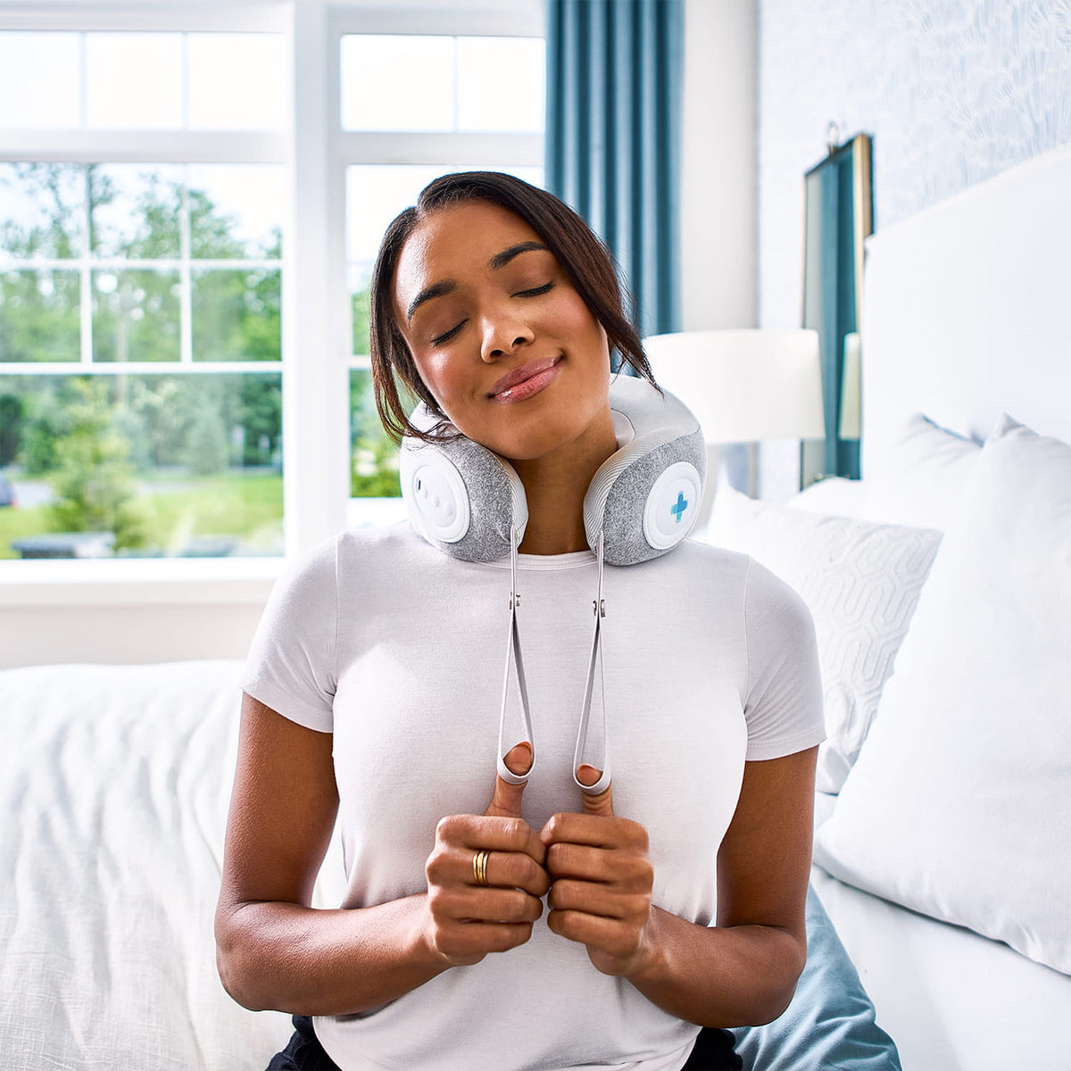 Woman relaxing with eyes closed, using MedMassager body massager on neck while lying on a sunlit bed.