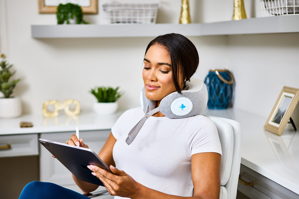 Woman using a MedMassager body massager on her neck while working at a desk, with shelves and plants in the background.