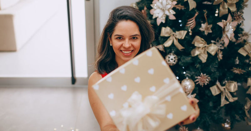 Smiling woman holding a Christmas gift in front of a decorated tree indoors.