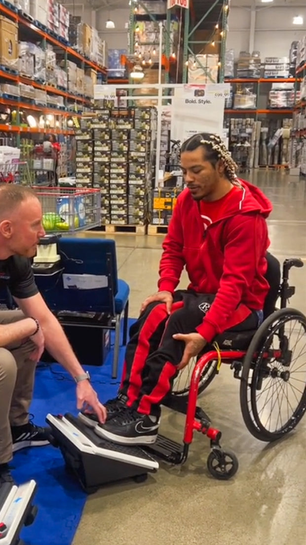 Man in wheelchair tries MedMassager foot massager with assistance from store worker in large warehouse store.