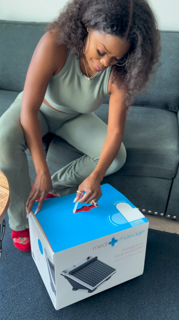 Woman opening MedMassager foot massager box while sitting on gray couch, wearing a green outfit.