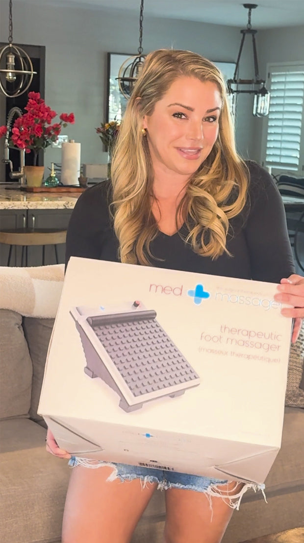 Woman sitting on a couch holding a MedMassager foot massager box in a modern living room.