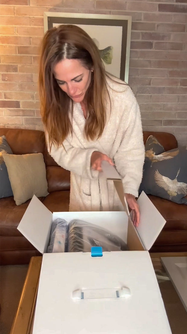 Woman in robe opens a MedMassager box on table in cozy living room with bird-print pillows.