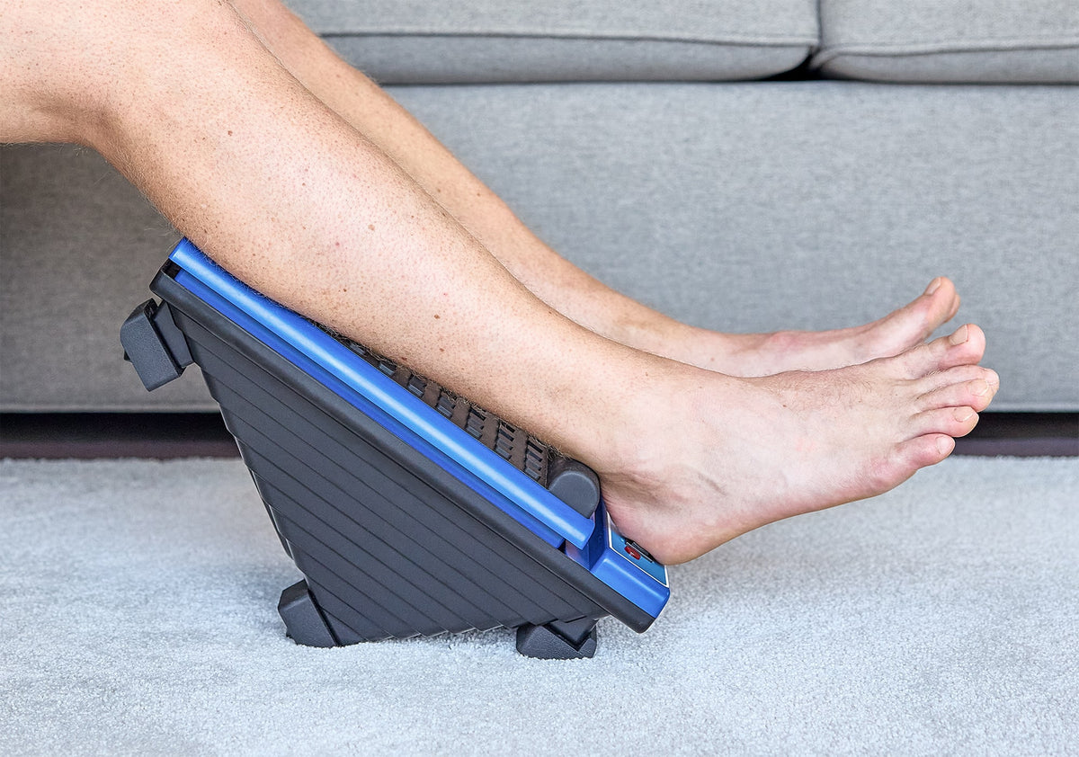 A person resting their bare feet on a black and blue ergonomic foot massager on a carpeted floor near a gray sofa.