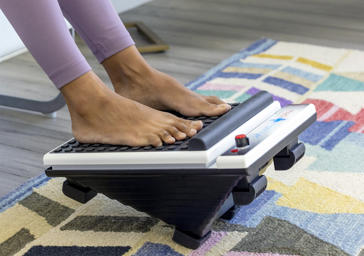 Lavender-legging wearer using MedMassager foot massager on a colorful rug.