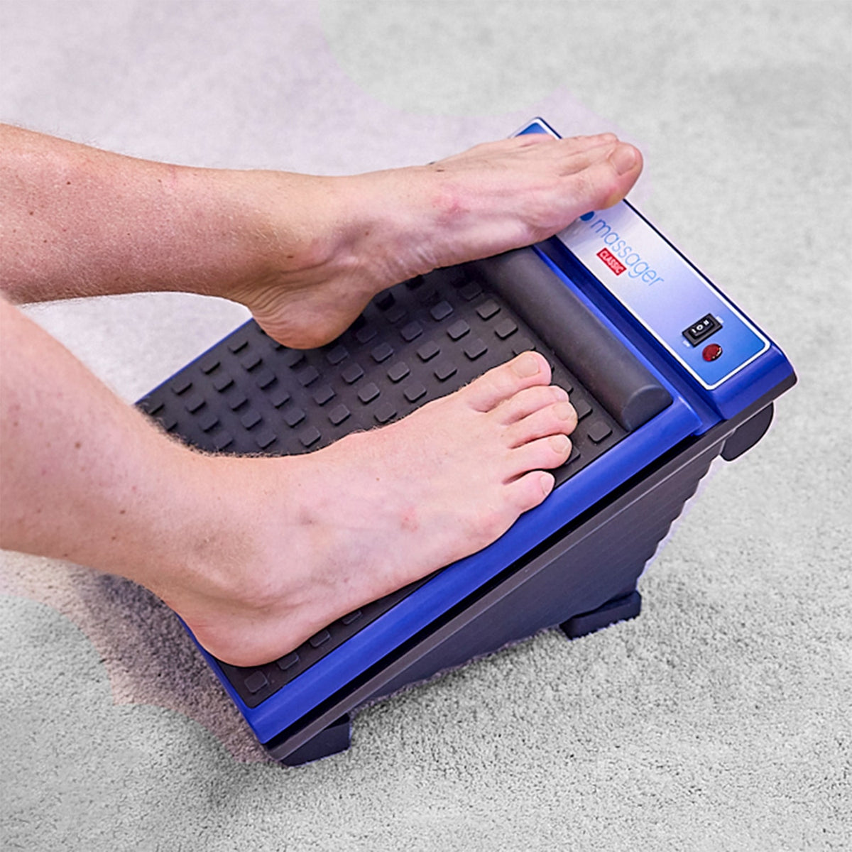 Feet using a MedMassager foot massager on a light-colored carpet.