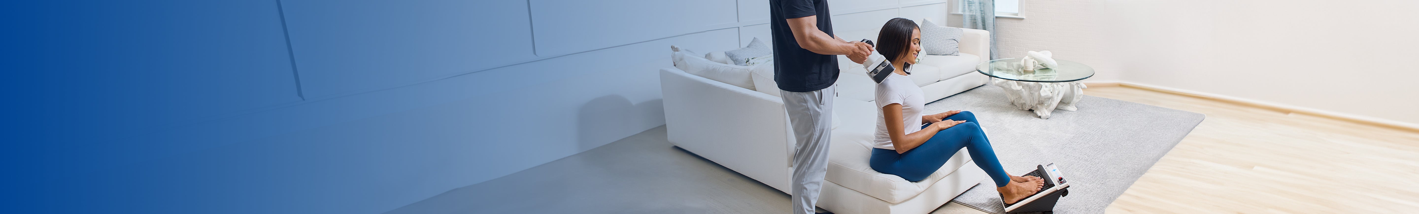 Man uses MedMassager body massager on woman’s shoulders as she sits on floor with tablet in a bright, modern living room.