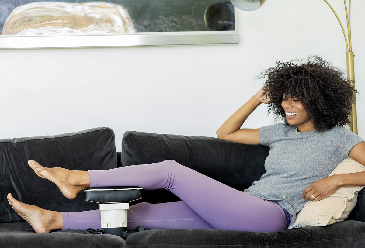 Woman using foot massager while relaxing on a couch, wearing a gray shirt and purple leggings.