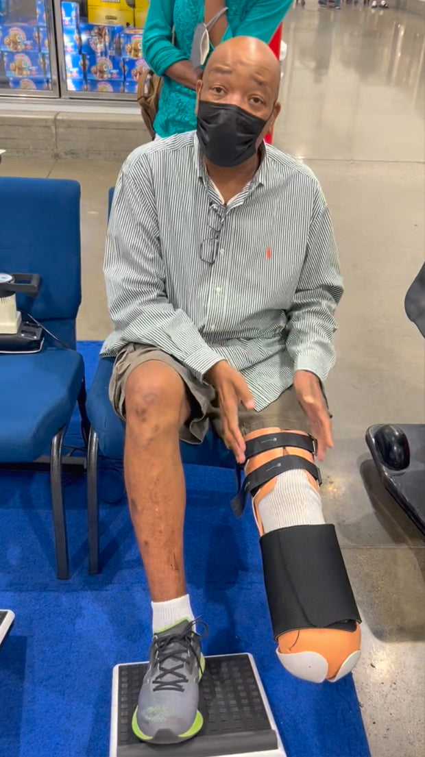 Man with prosthetic leg sits on chair in public space, wearing mask and using MedMassager foot massager.