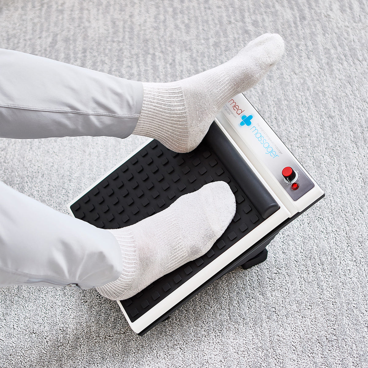 Person using MedMassager foot massager on light gray carpet, wearing white socks, for plantar fasciitis relief.