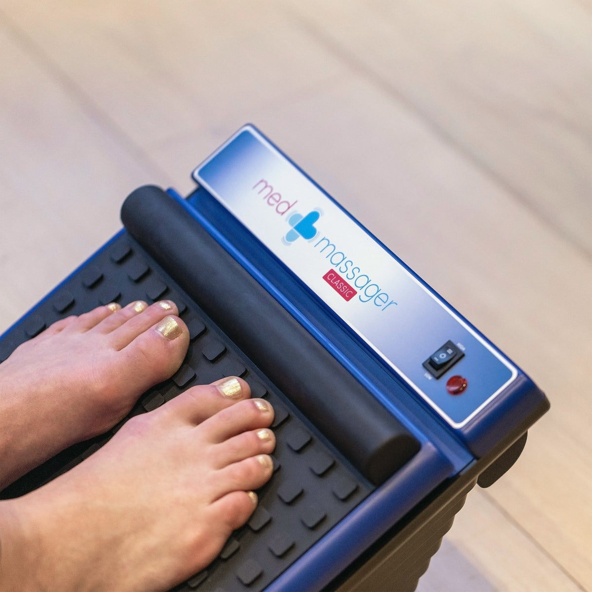 Feet using a MedMassager foot massager on a wooden floor.