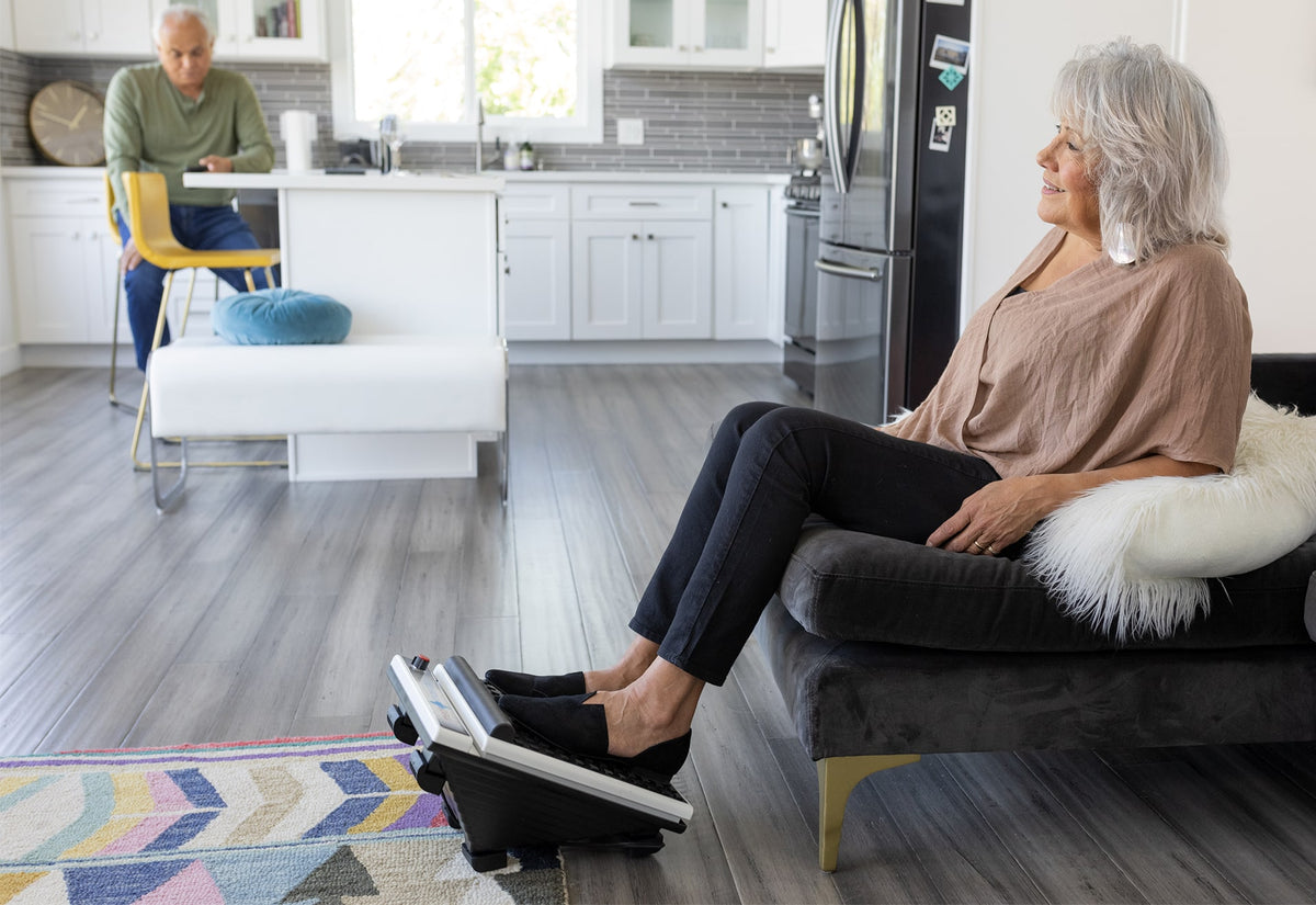 Older woman using foot massager in living room, relaxing while a man sits at the kitchen counter in the background.