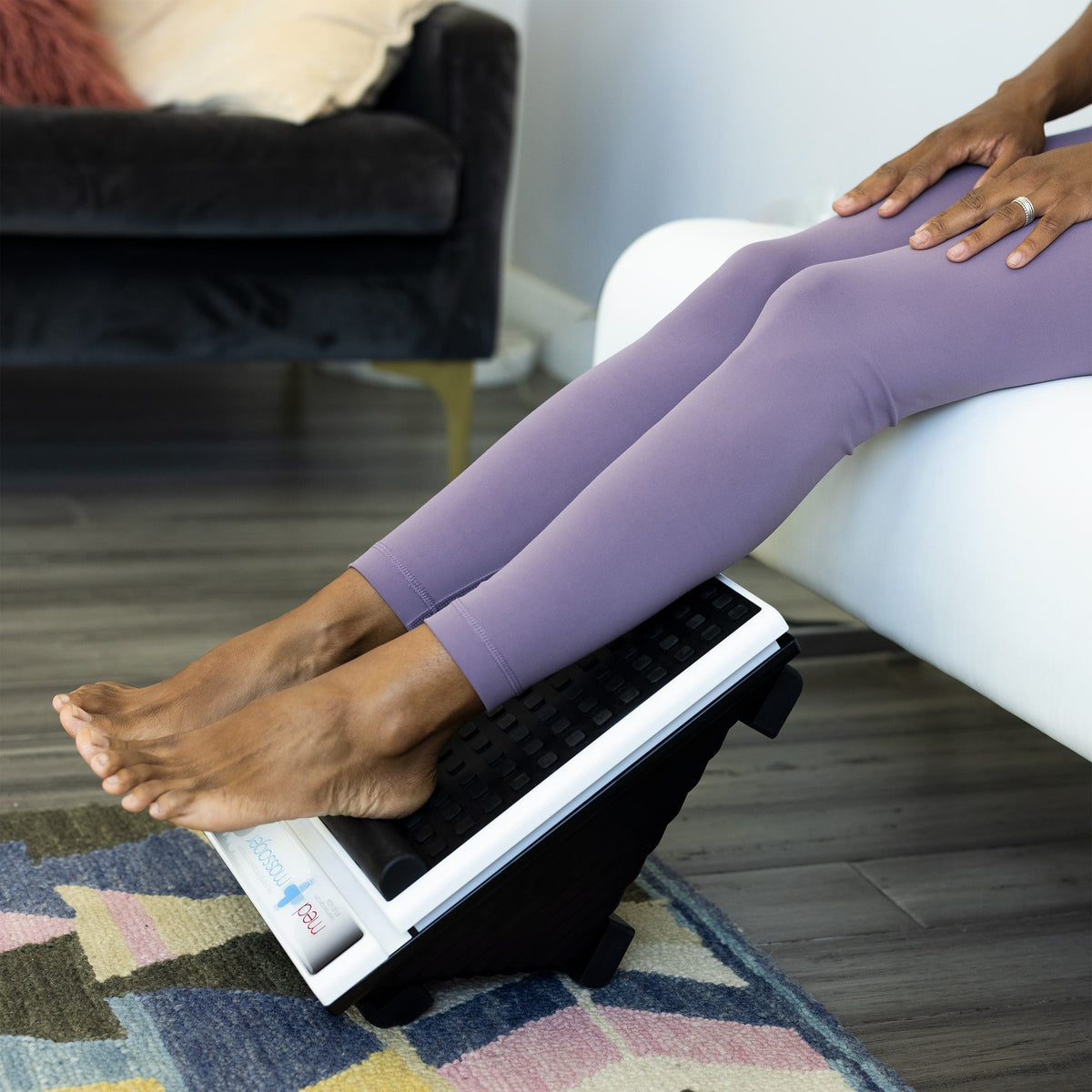 Feet resting on MedMassager foot massager, black wedge-shaped device, on a white couch.