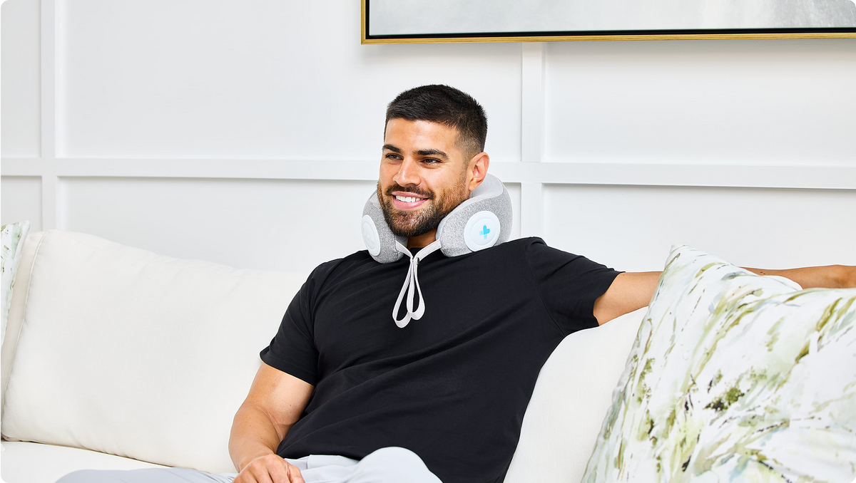 Man sitting on a white couch, smiling, using a gray MedMassager body massager with controls in a bright modern room.