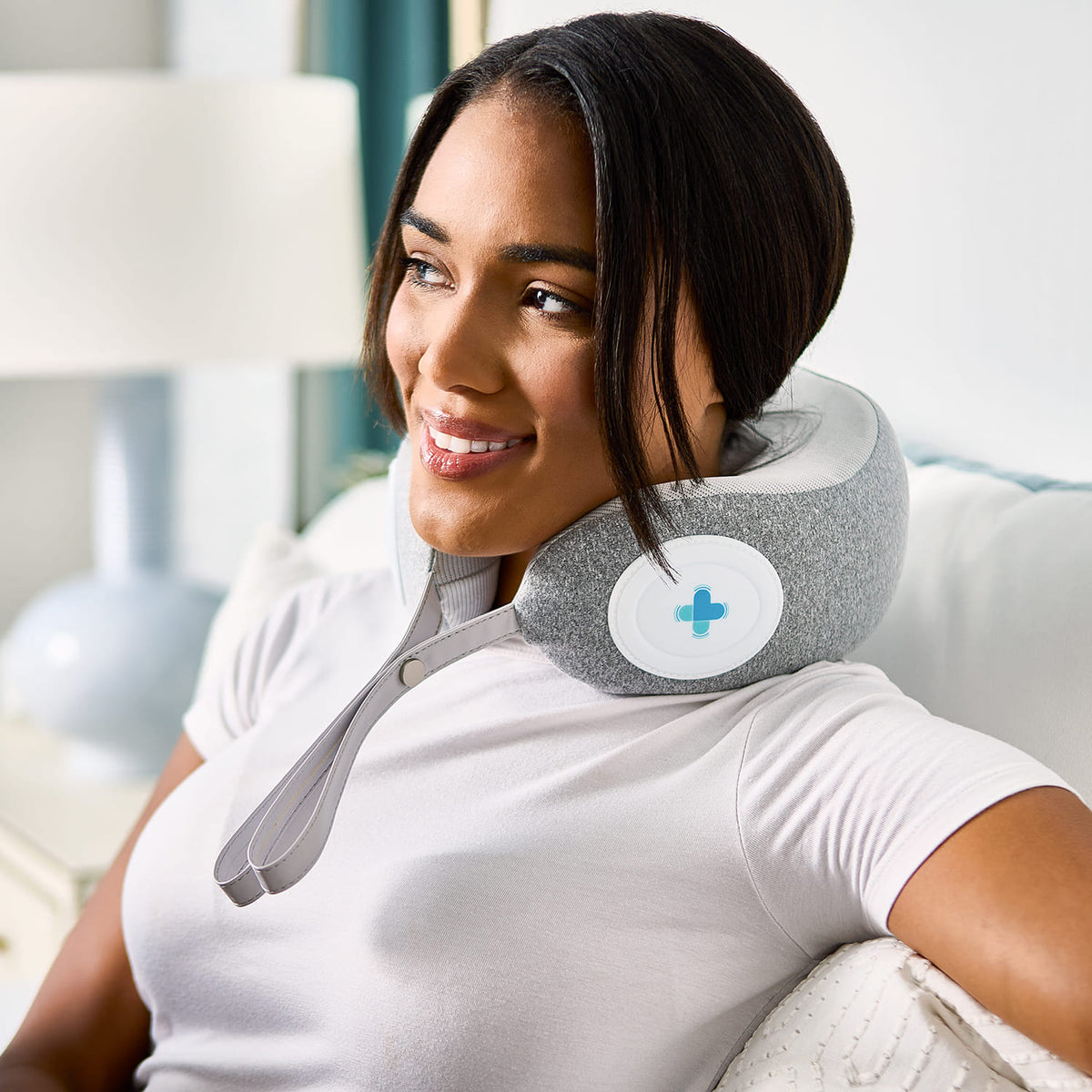 Woman uses MedMassager body massager on her neck, smiling and relaxing on a couch for pain relief.